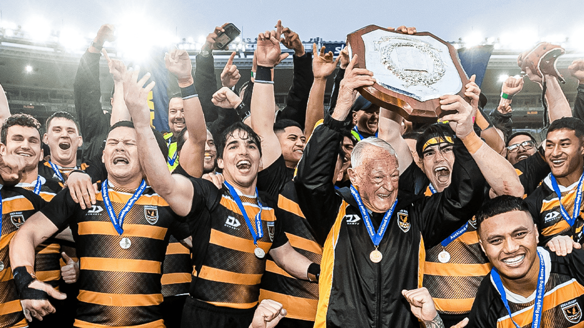 Auckland Rugby club team celebrating a championship victory on the field, lifting the Gallaher Shield trophy in front of a cheering crowd.
