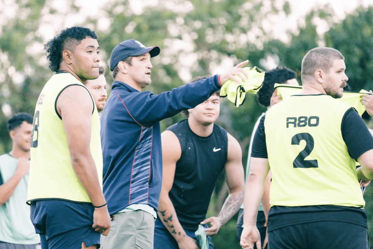 Coach directing players during an Auckland Rugby training session on a grass field, with athletes wearing training bibs and listening to instructions.